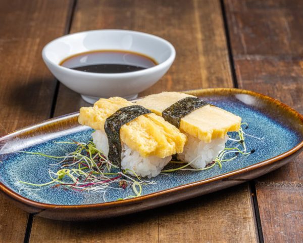 Nigiri-omelette-japonaise-tamago Nigiri délicatement préparé, surmonté d'une tranche d'omelette japonaise tamago moelleuse et dorée, reposant sur un lit de riz vinaigré.